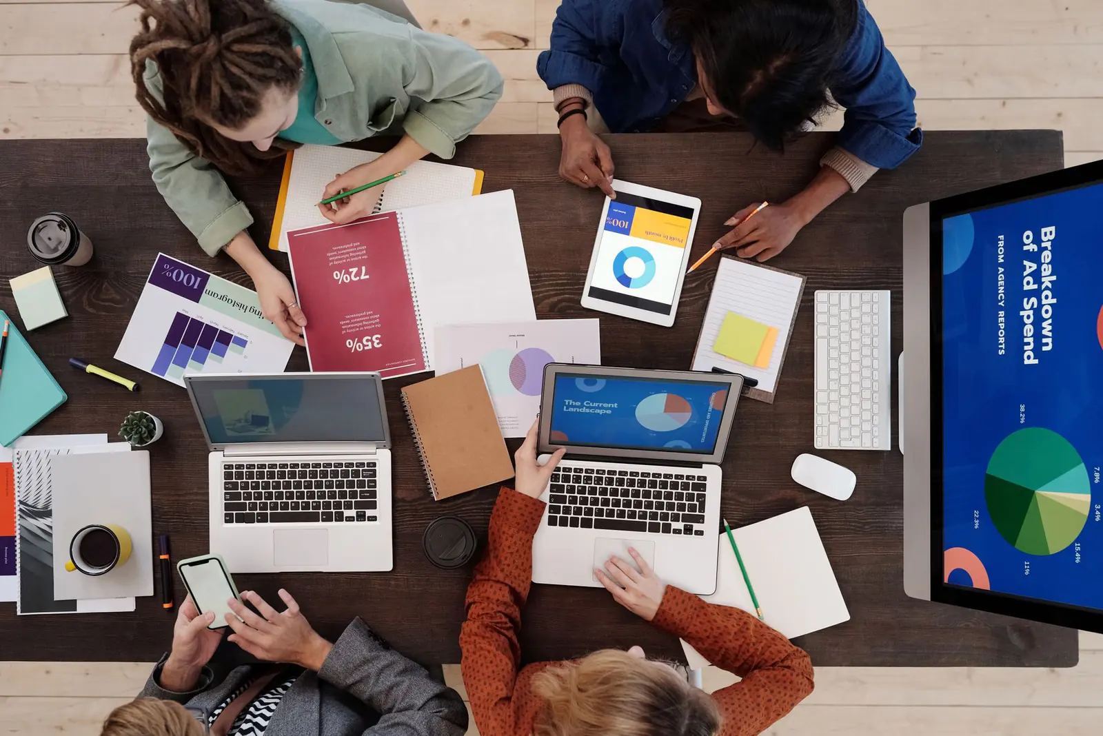 Four people working at a desk covered in laptops, charts and notes, seen from above